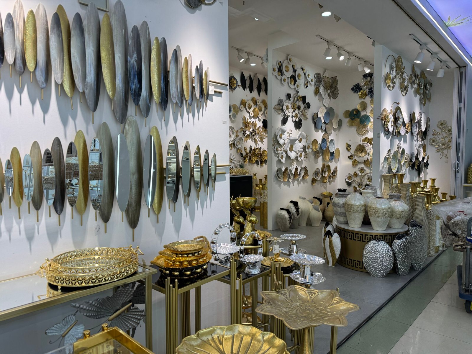 Interior of a Yiwu home decor market shop with rows of oval metal wall art panels, round mirrors, gold trays and decorative ceramic vases on display.