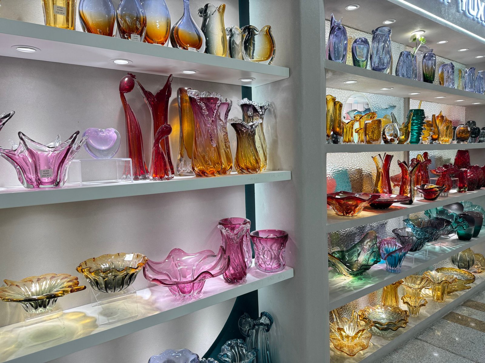 Pink, purple, amber and multicolor art glass vases and sculptural bowls filling lit display shelves in a wholesale glass decor shop at Yiwu home decor market.