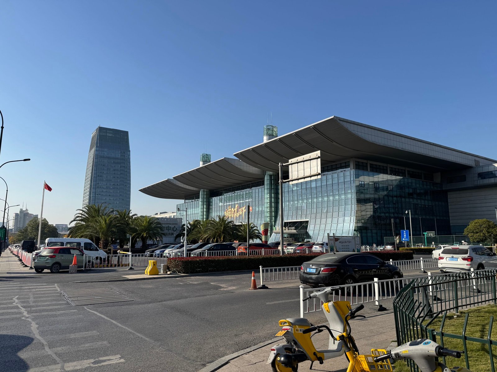 Exterior of Yiwu International Trade City District 4 South Gate in Yiwu, Zhejiang, China, with modern glass-fronted building, curved roof, palm trees, Chinese flag and parked cars on a sunny day.