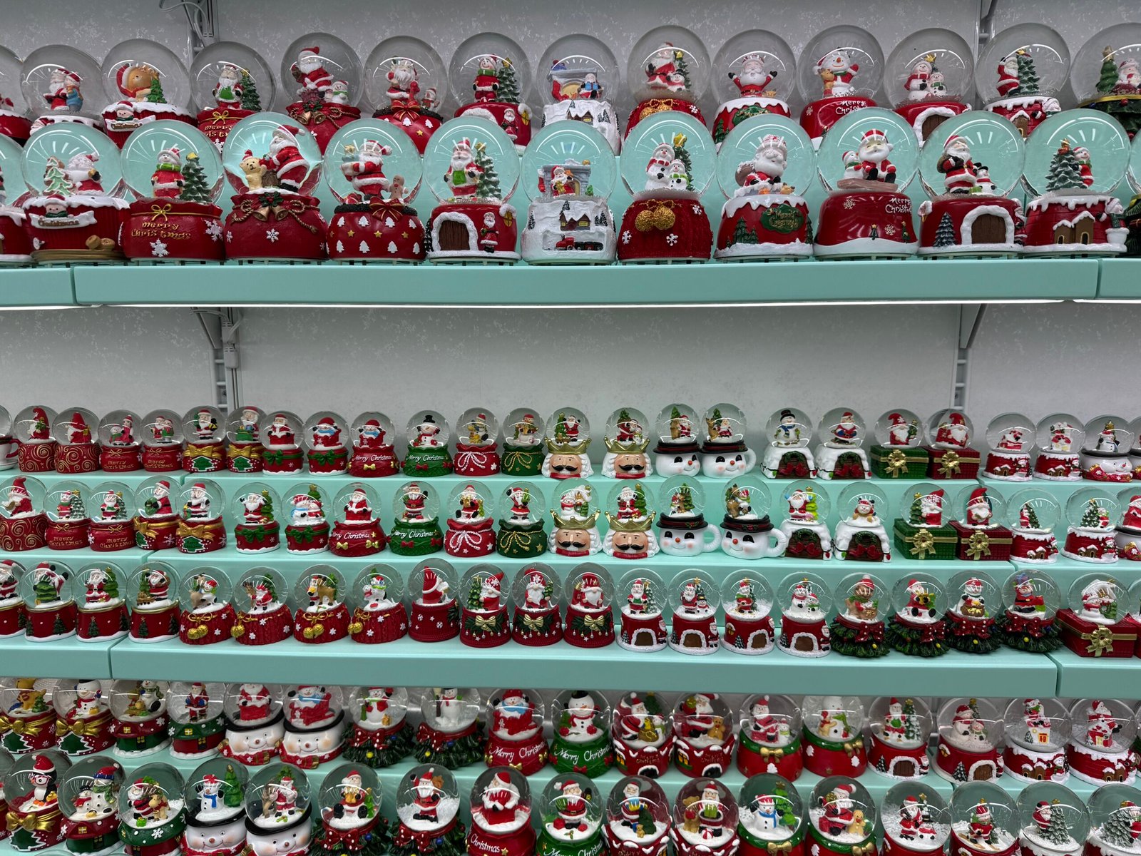 Multiple rows of mini Christmas snow globes with Santa and snowman scenes displayed on turquoise shelves in a Yiwu shop.