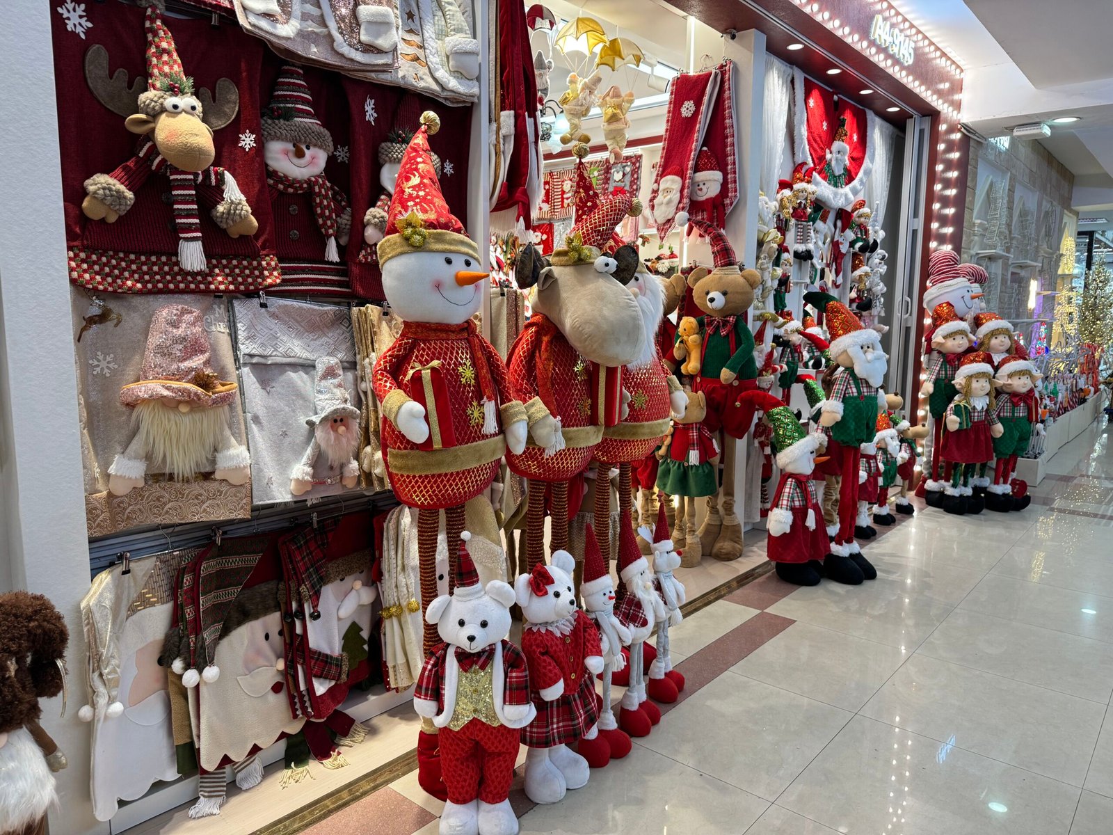 Plush snowmen, reindeer and teddy bear dolls standing at the entrance of a Yiwu Christmas wholesale shop with textile wall hangings behind them.