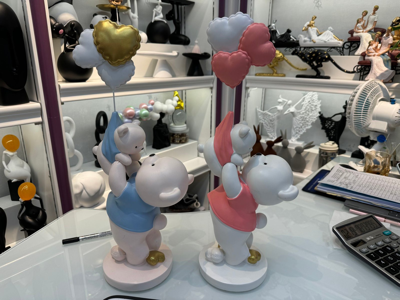 Two resin bear parent and baby figurines in blue and pink outfits holding heart-shaped balloons on a white table inside a resin crafts shop at the Yiwu home decor market in China.