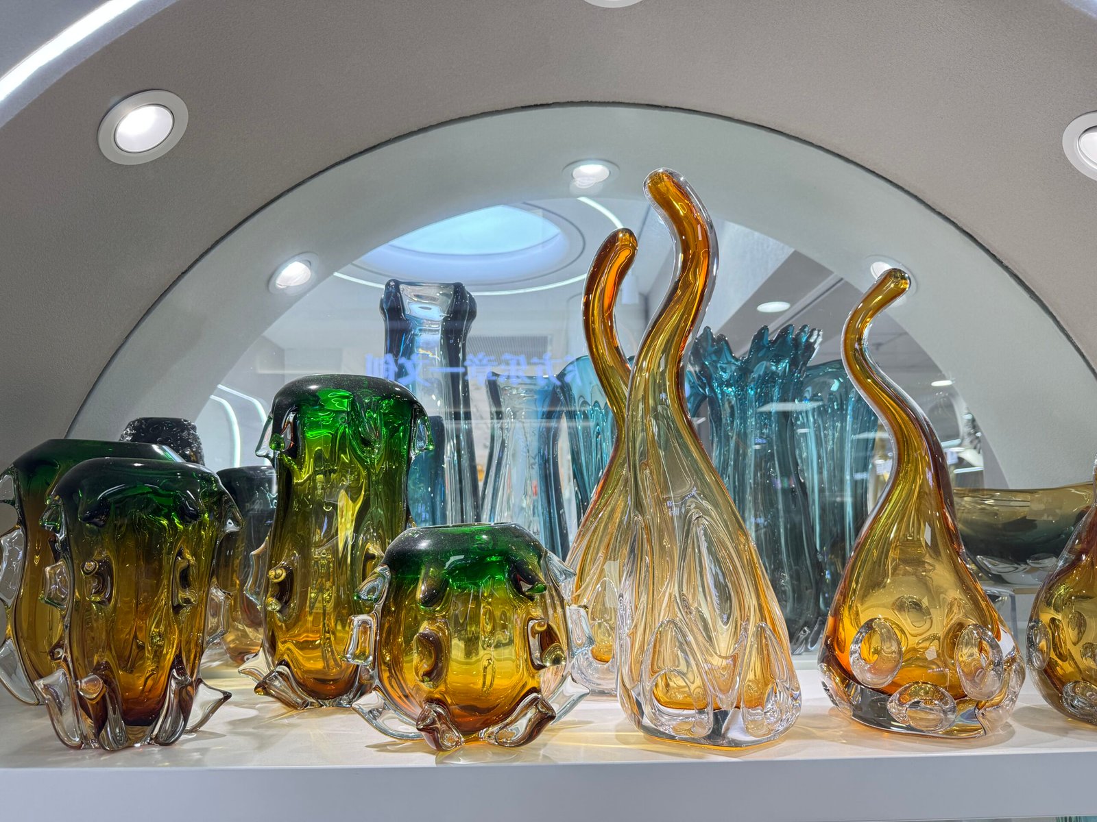 Green and amber sculptural art glass vases and flame-shaped pieces under an arch display in the Yiwu home decor market.