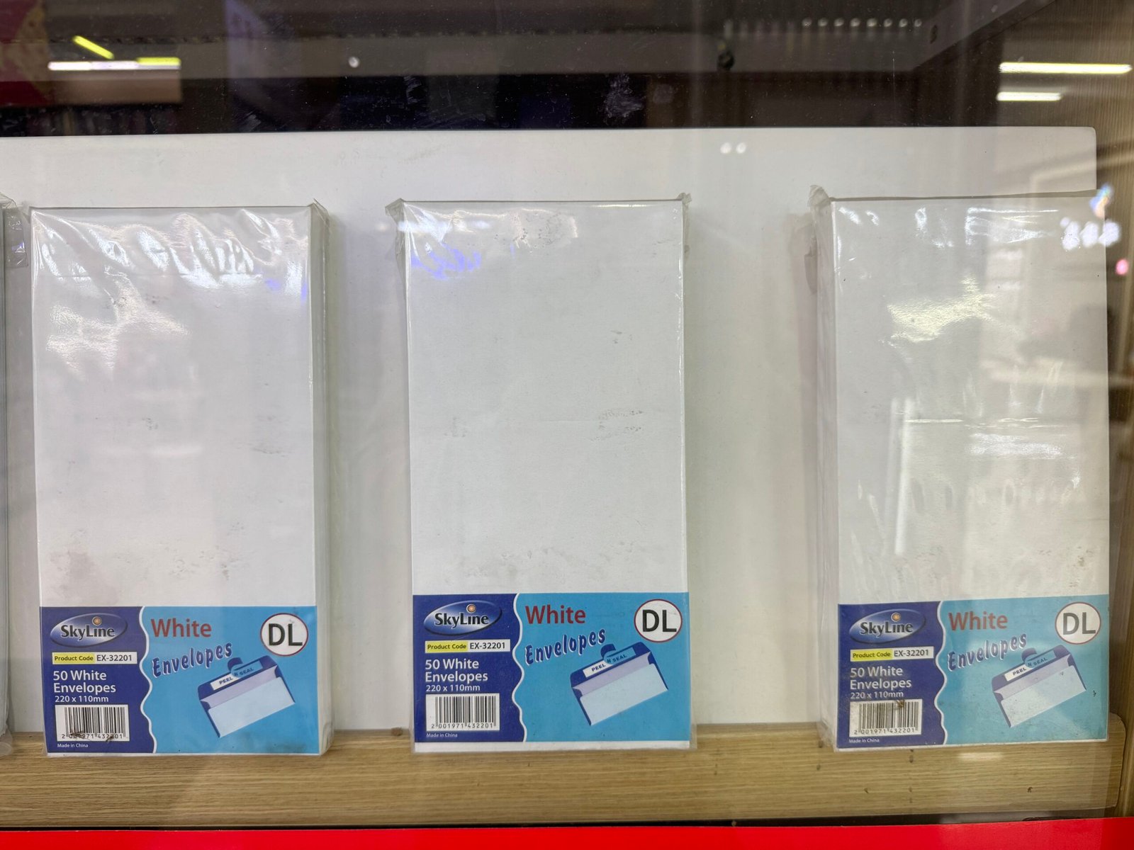 Three packs of DL white envelopes 220×110mm, 50pcs per pack, standing on a wooden shelf at a Yiwu International Trade City stationery shop.
