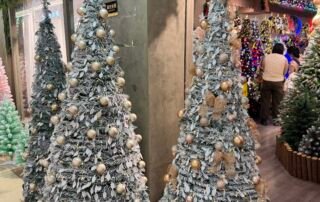 Two snow-covered artificial Christmas trees with gold baubles and bows at the corner of a Yiwu Christmas decorations shop.