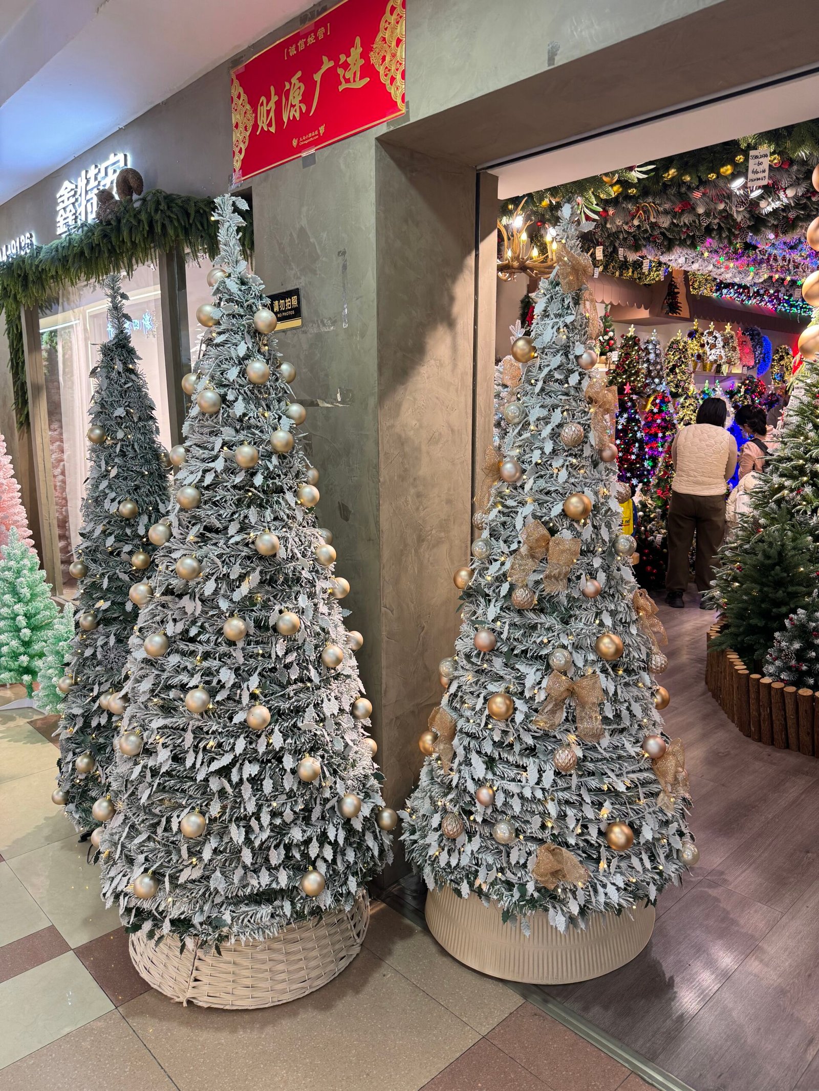 Two snow-covered artificial Christmas trees with gold baubles and bows at the corner of a Yiwu Christmas decorations shop.