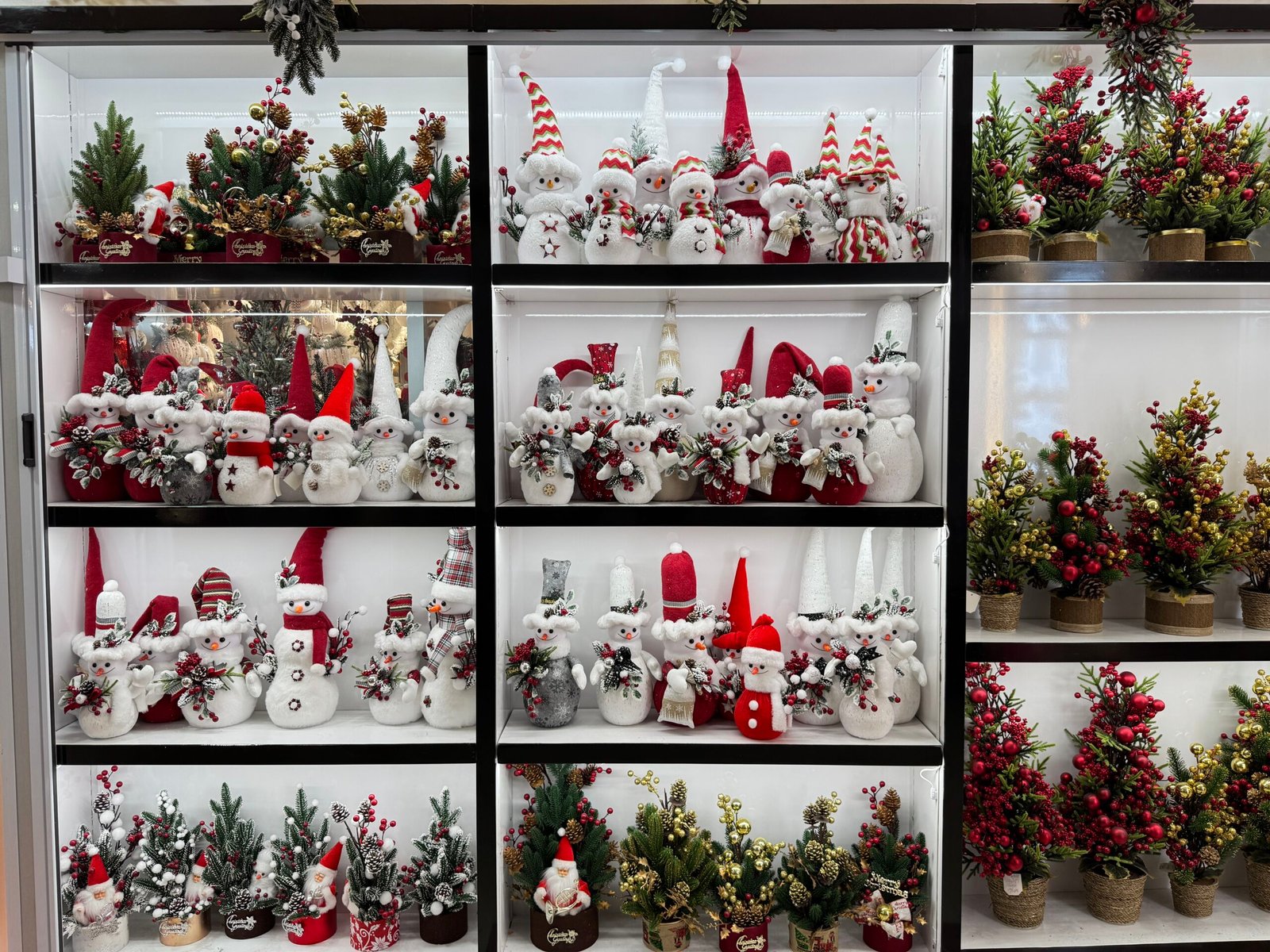 White snowman and Santa plush ornaments with small potted berry trees arranged on shelves at Yiwu Christmas wholesale market.