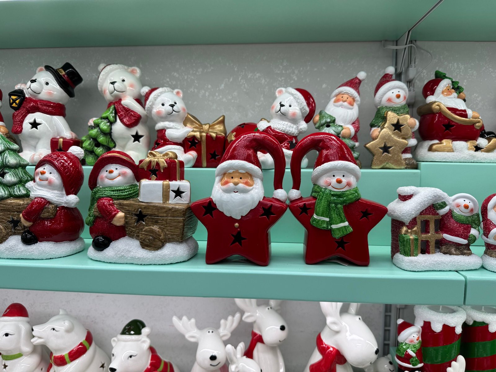 Close-up image of brightly painted ceramic Christmas figurines on a shelf in Yiwu, China, showing polar bears in red scarves holding trees and gifts, jolly snowmen, Santas riding sledges and two large red star-shaped lantern figures with Santa and a snowman in green scarf.