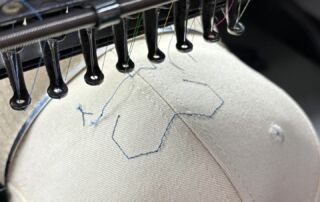 Embroidery machine stitching a placement outline on a beige baseball cap mounted on a cap frame.