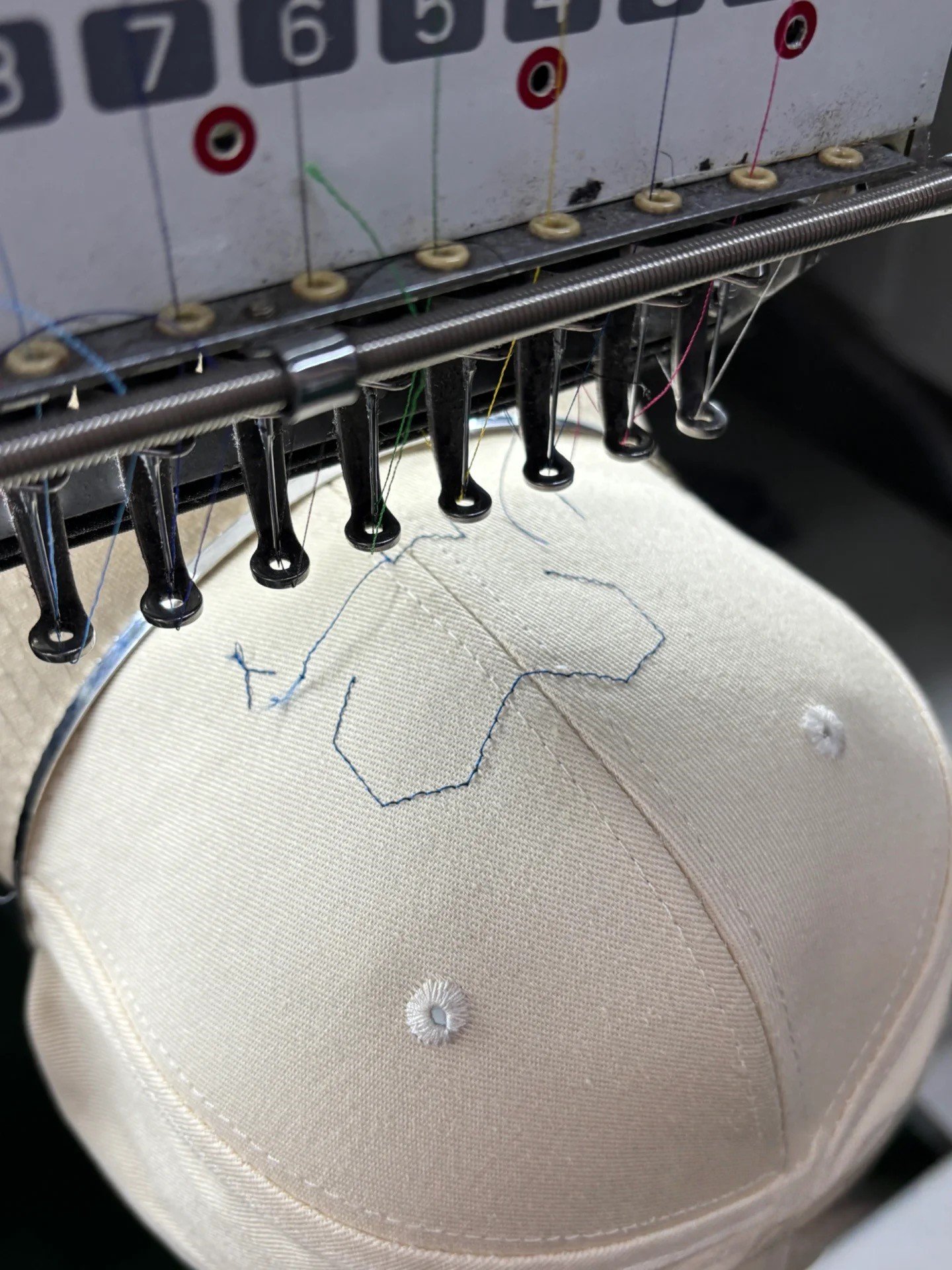 Embroidery machine stitching a placement outline on a beige baseball cap mounted on a cap frame.