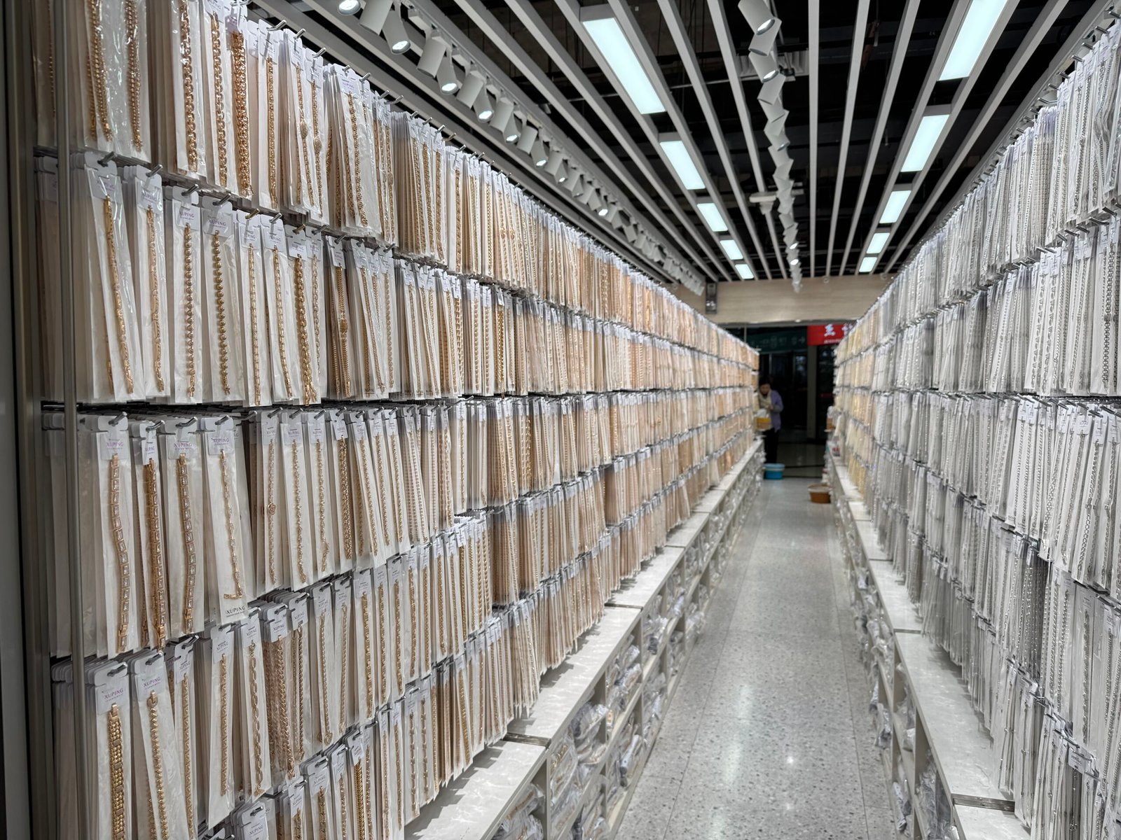 Long narrow aisle with dense rows of packaged chain necklaces in gold and silver tones on both sides in a Yiwu jewelry wholesale market.