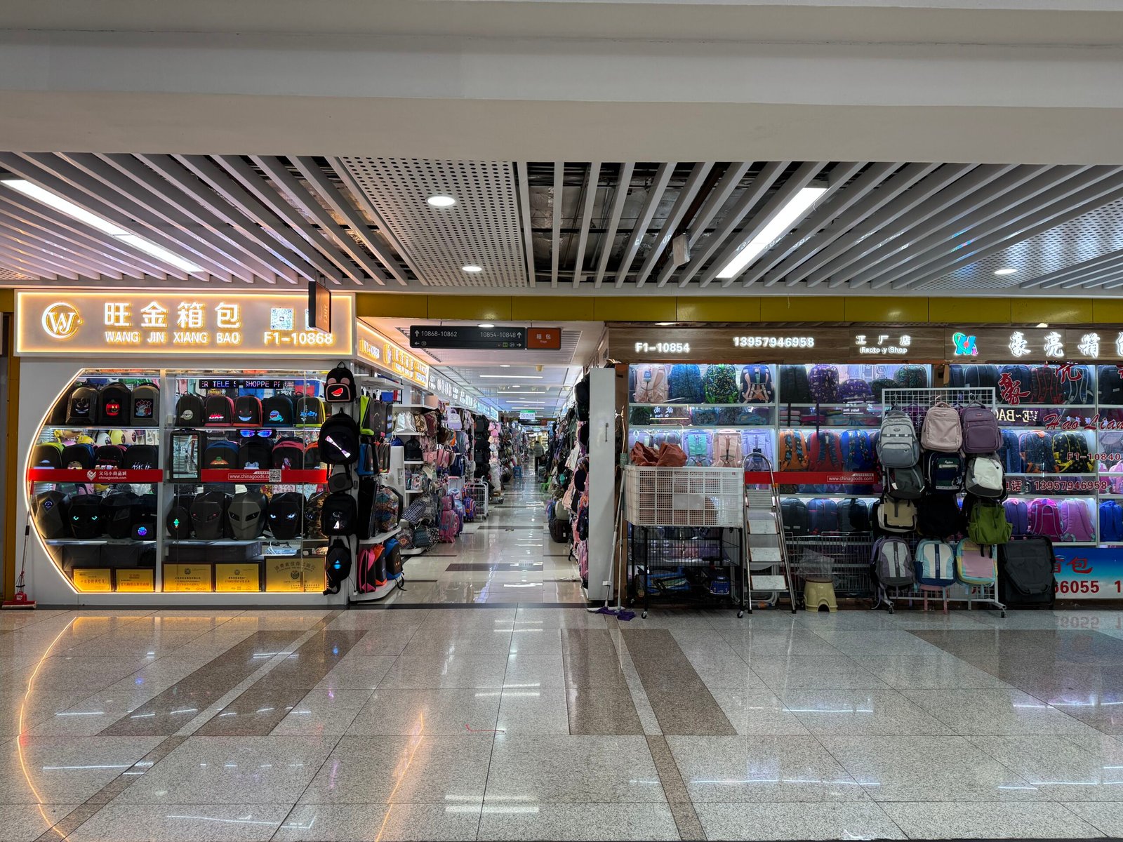 Backpack wholesale shops in Yiwu Bags & Luggage Wholesale Market, with illuminated storefront signs and rows of backpacks and schoolbags along the main aisle.