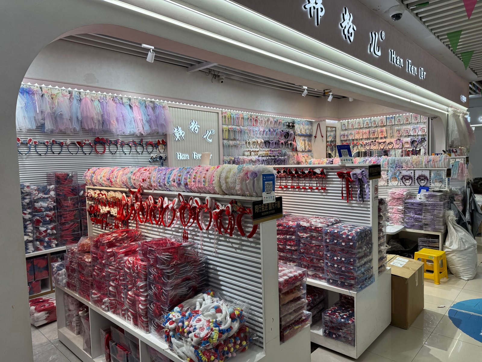 Yiwu hair accessories wholesale booth with rows of headbands, hair bows, scrunchies and packaged hair clips on white display racks, Yiwu hair items wholesale market