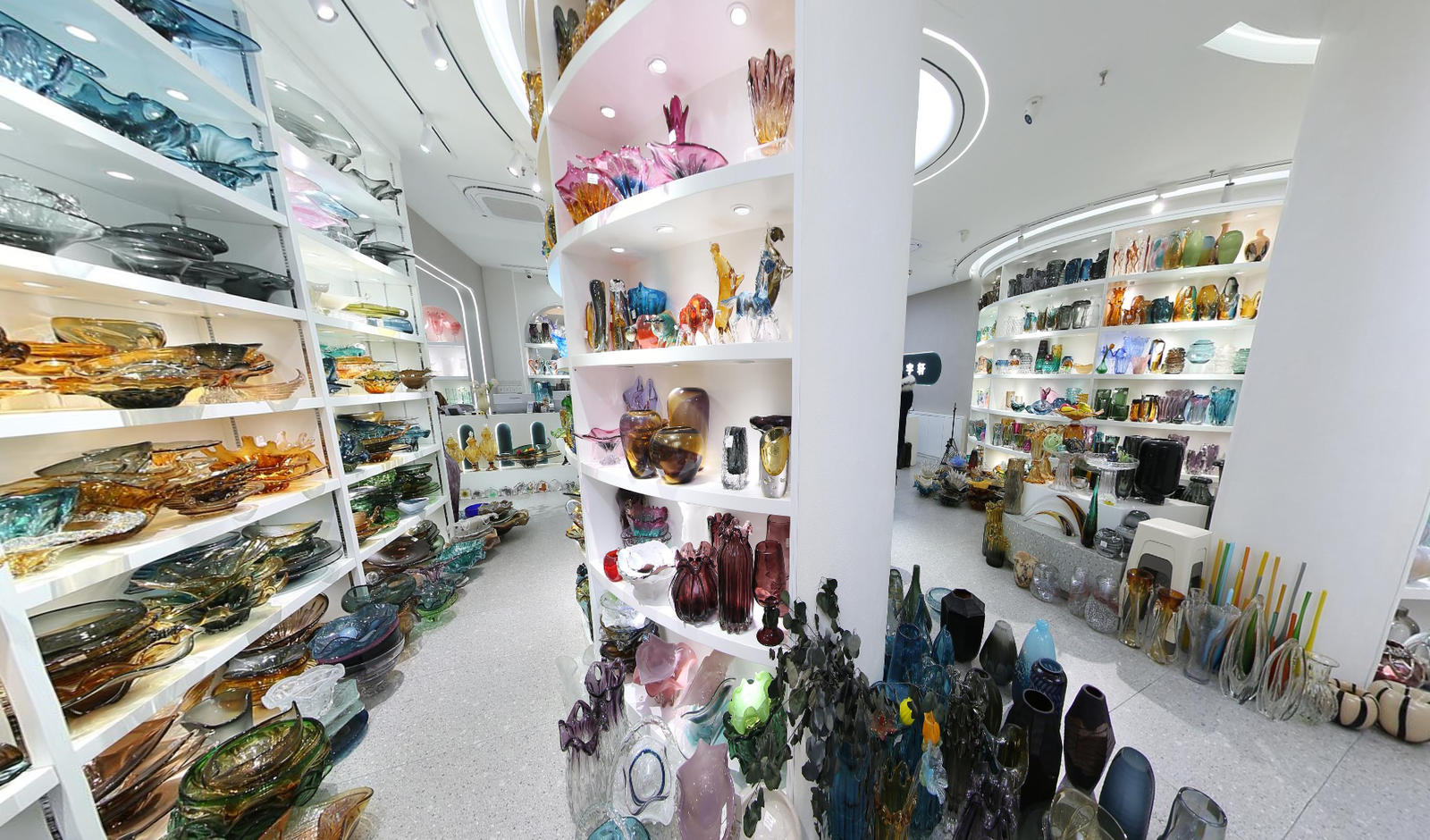 Wide-angle view inside a Yiwu glass decor showroom with a curved display island, wall shelves of glass plates and bowls, and colorful decorative glass vases.