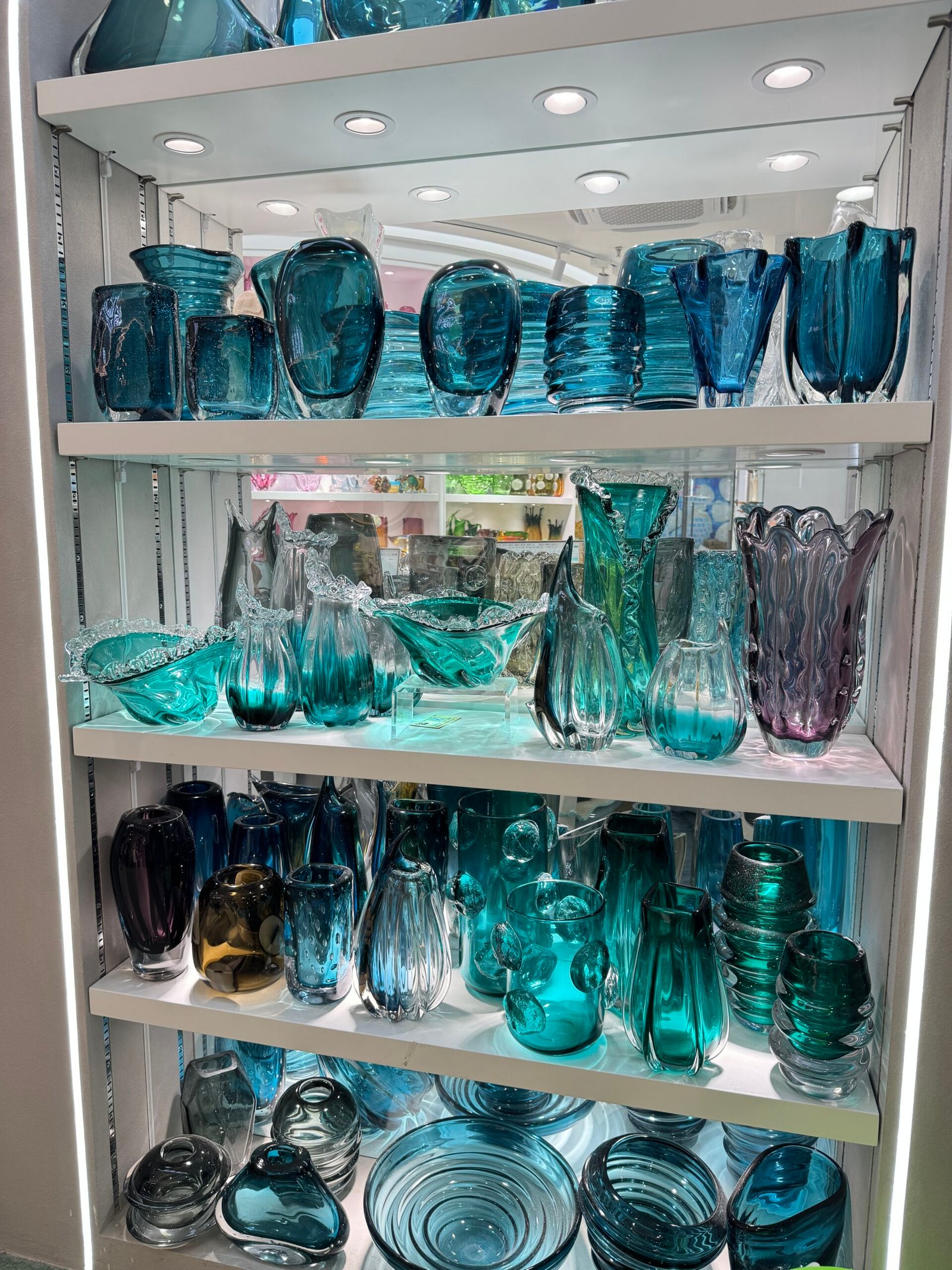 Teal and turquoise glass decor display shelf with modern glass vases, bowls and plates under spotlights in a Yiwu showroom.