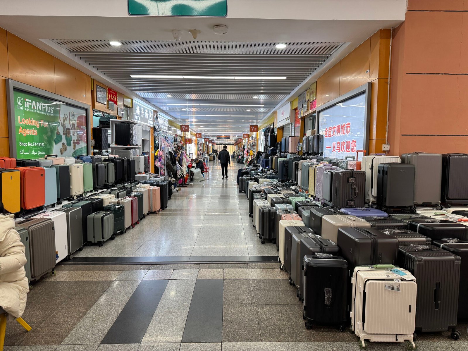 Luggage wholesale aisle in Yiwu market with rows of hard-shell suitcases and carry-on trolley cases displayed along both sides of the corridor.