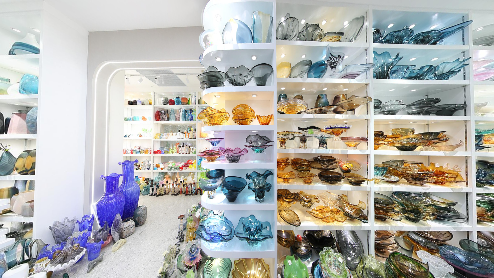 Wide view of a bright Yiwu glass decor showroom with white wall shelves filled with blue, amber and smoke glass bowls, plates and decorative vases.