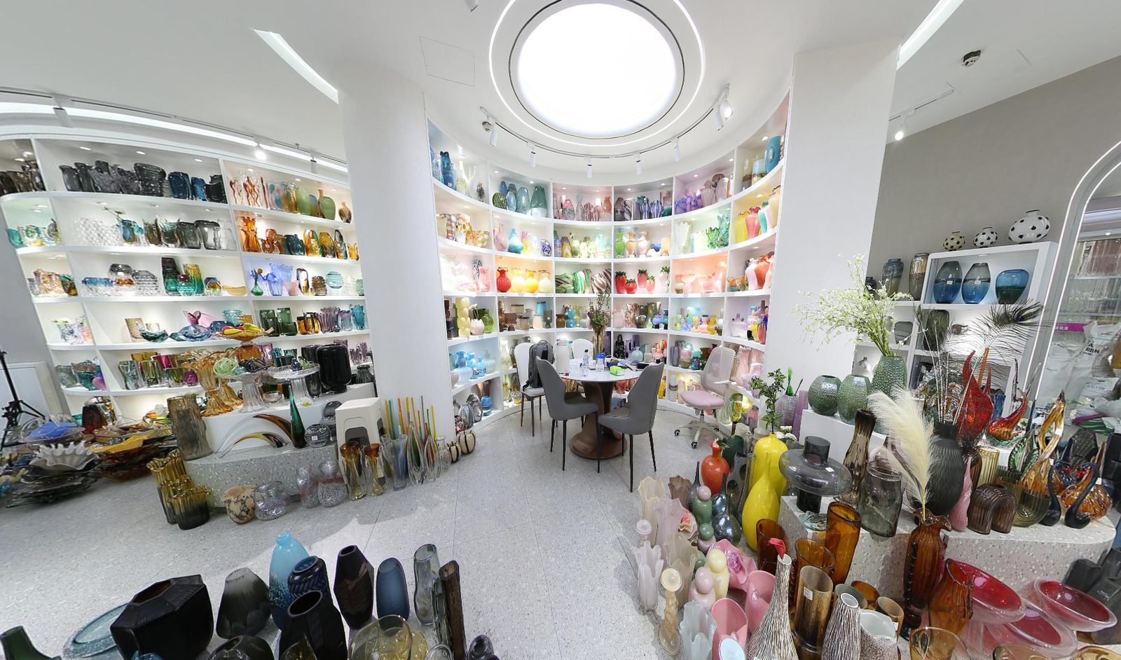 Round showroom area in Yiwu market with a meeting table and curved wall shelves full of colorful glass vases, bowls and decorative glass items.