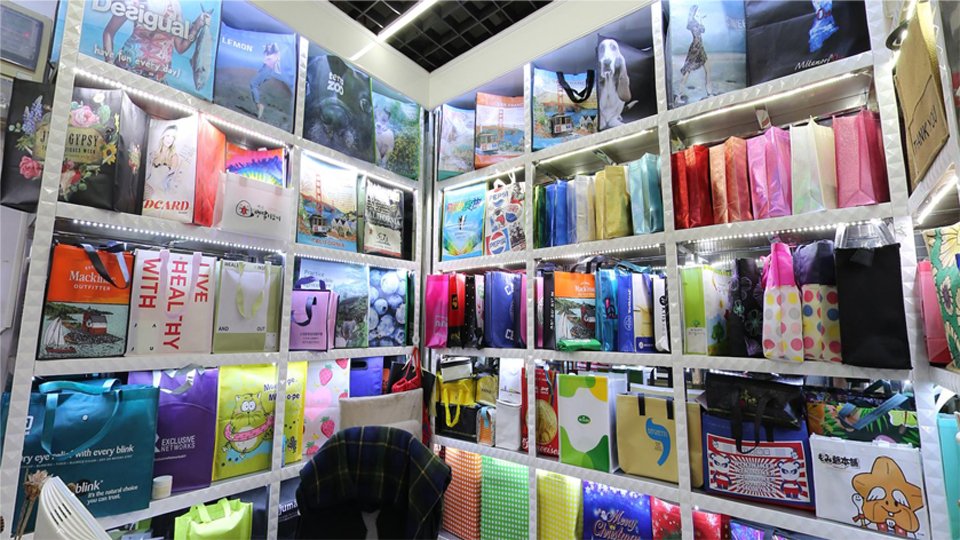 Sample display of custom non-woven shopping bags in Yiwu Market showing various sizes, colors, printed logos, laminated finishes, and handle styles from a bag manufacturer