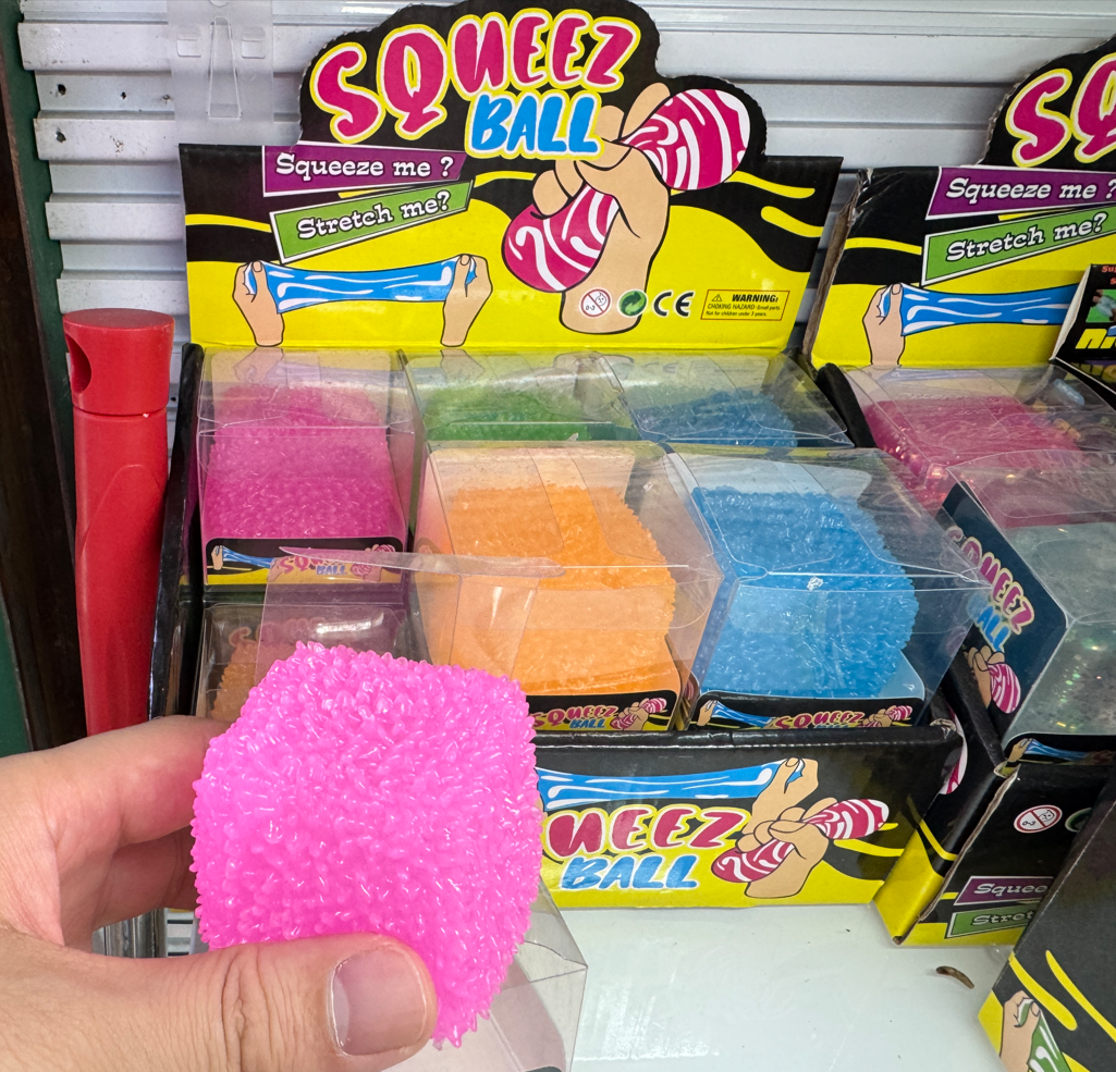 Bright pink textured squeeze ball stress relief toy with a soft bumpy surface, held in hand in front of assorted wholesale squeeze ball display boxes at Yiwu Toy Market.