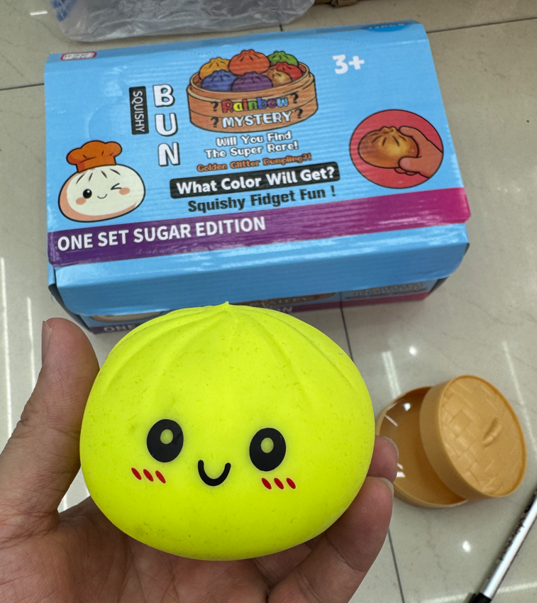 Cute yellow squishy bun fidget toy with a smiling face, soft stress relief texture, capsule container, and blue mystery box packaging, photographed at Yiwu Toy Market for wholesale sensory toy buyers.